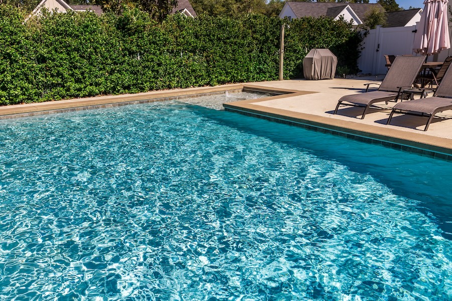 A rectangular new swimming pool with tan concrete edges in the fenced backyard of a new construction house with privacy hedges.