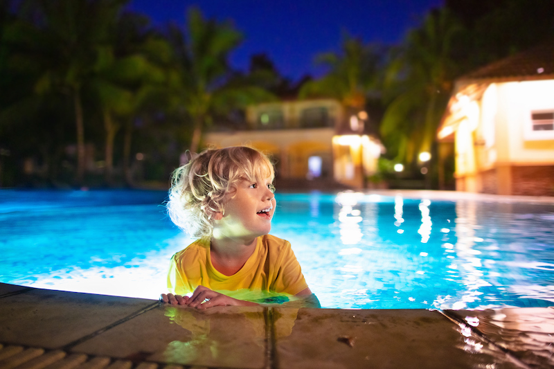Kids play in outdoor swimming pool of tropical resort at night. Evening swim on exotic island. Baby learning to dive. Children playing in water. Beach and summer fun.