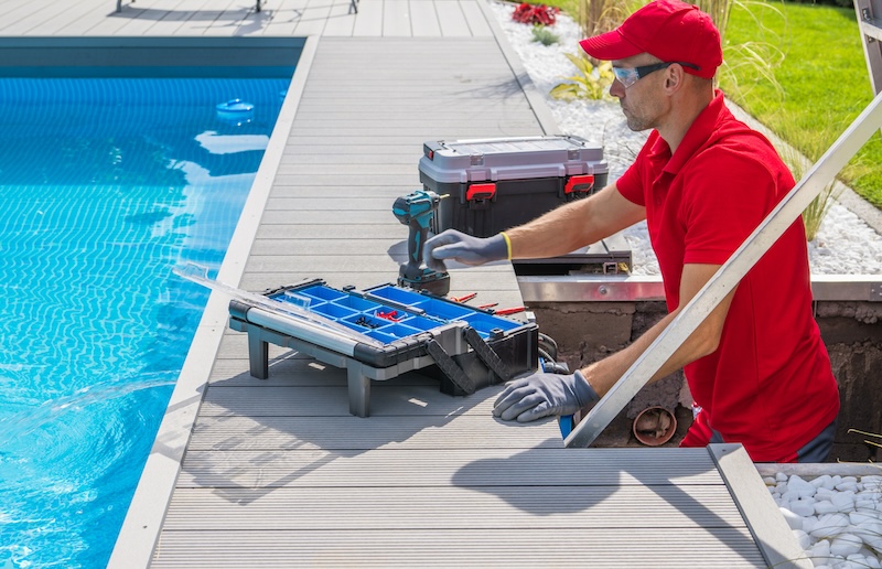 Outdoor Swimming Pool Heating and Filtering System Maintenance Performed by Caucasian Middle Aged Technician. Open Toolbox and Electric Drill Driver on the Floor in Front of Him.