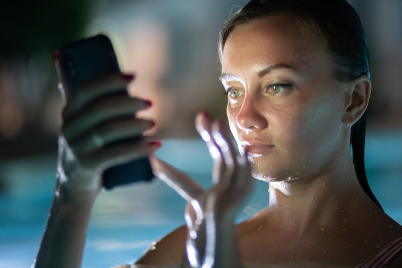 woman using mobile cell phone app on vacation in pool at night on travel holidays. Girl using smartphone app looking at screen smiling happy. Screen light on face.