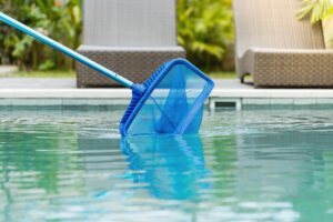 Cleaning swimming pool of fallen leaves with skimmer net equipment against sun resort loungers at sunny summer day