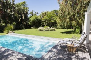 Sparkling pool with a lush backyard behind it. Two comfy lawn chairs sit on the right side of the pool deck.