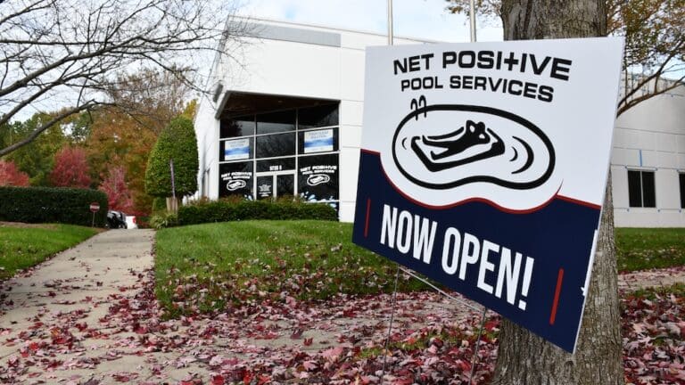 Outside view of the Net Positive Pool Store. A sign showing the logo and text that reads "now open" is in the forefront of the picture with the store entrance in the back.