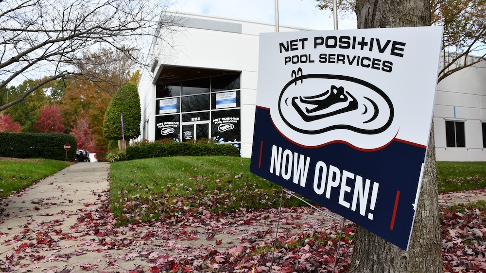 Outside view of the Net Positive Pool Store. A sign showing the logo and text that reads "now open" is in the forefront of the picture with the store entrance in the back. 