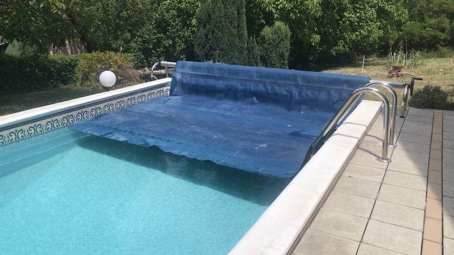 Angled perspective view of half removed blue solar pool cover with clean swimming pool water and stainless steel ladder