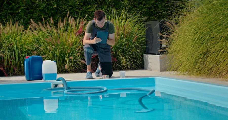 Pool technician kneeling by the pool, using a tablet to check water quality. Surrounded by chemical containers and lush greenery, highlighting the importance of professional maintenance in a clean, outdoor setting.