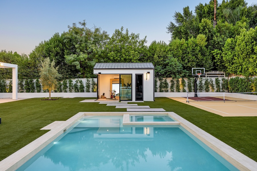 Modern backyard with a pool, basketball court, and garden house surrounded by lush greenery under a clear sky.