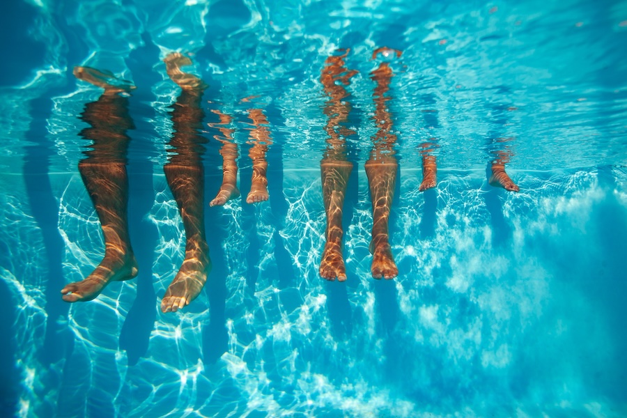 Underwater view of diverse people's legs, adults and kids, suspended in a clear blue sunny swimming pool dangling around