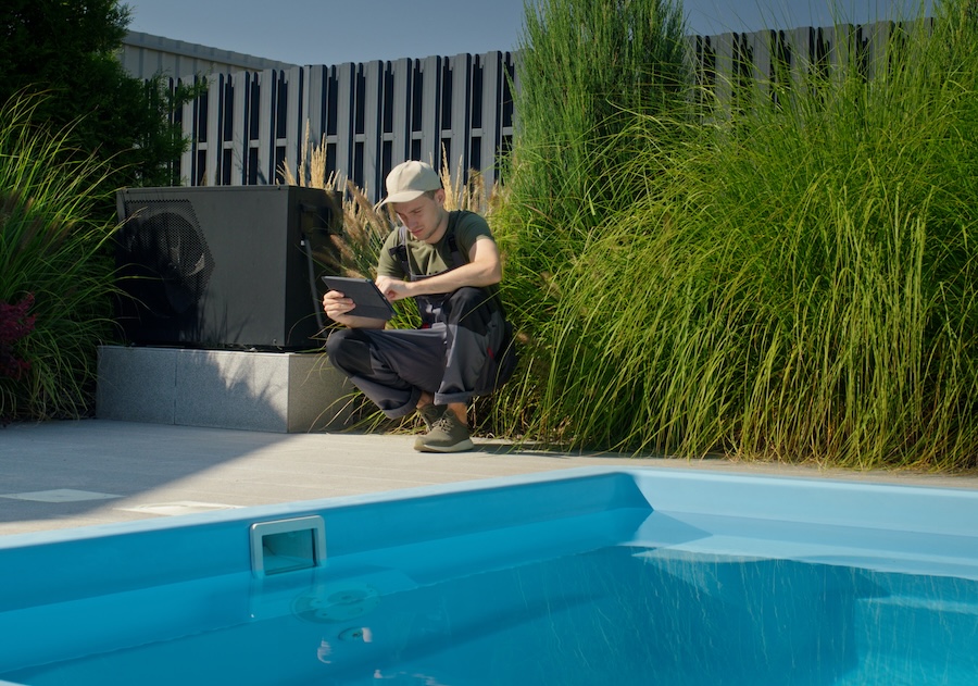 A technician is crouched by a pool, intently focused on the tablet in his hands. He is surrounded by lush green plants, contrasting with the bright blue of the pool. The setting suggests a maintenance or repair task being conducted outdoors, blending technology with the natural environment. The scene captures a moment of work in a peaceful and well-maintained space, emphasizing the blend of nature and modernity.
