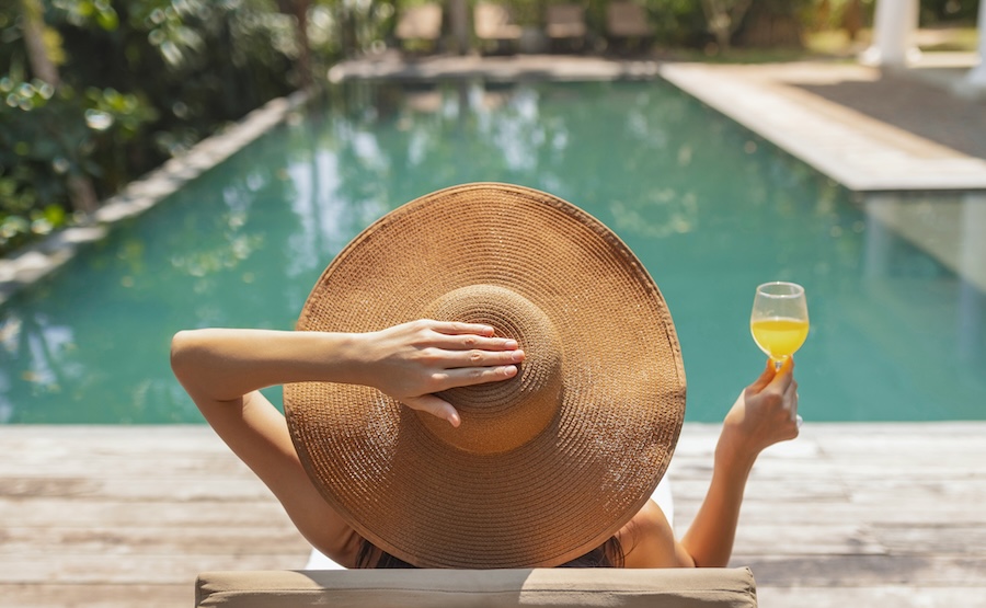 A woman wearing a large sun hat sits by the pool with a refreshing drink in hand. The tropical surroundings and calm atmosphere offer a perfect spot for relaxation and enjoyment.