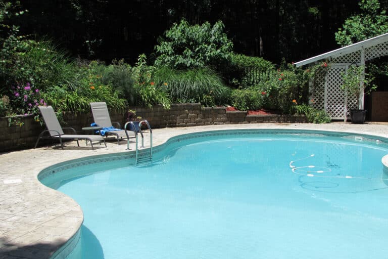 In-ground backyard swimming pool with clear blue water surrounded by a concrete patio with outdoor lounge chairs and a white lattice shed in the background